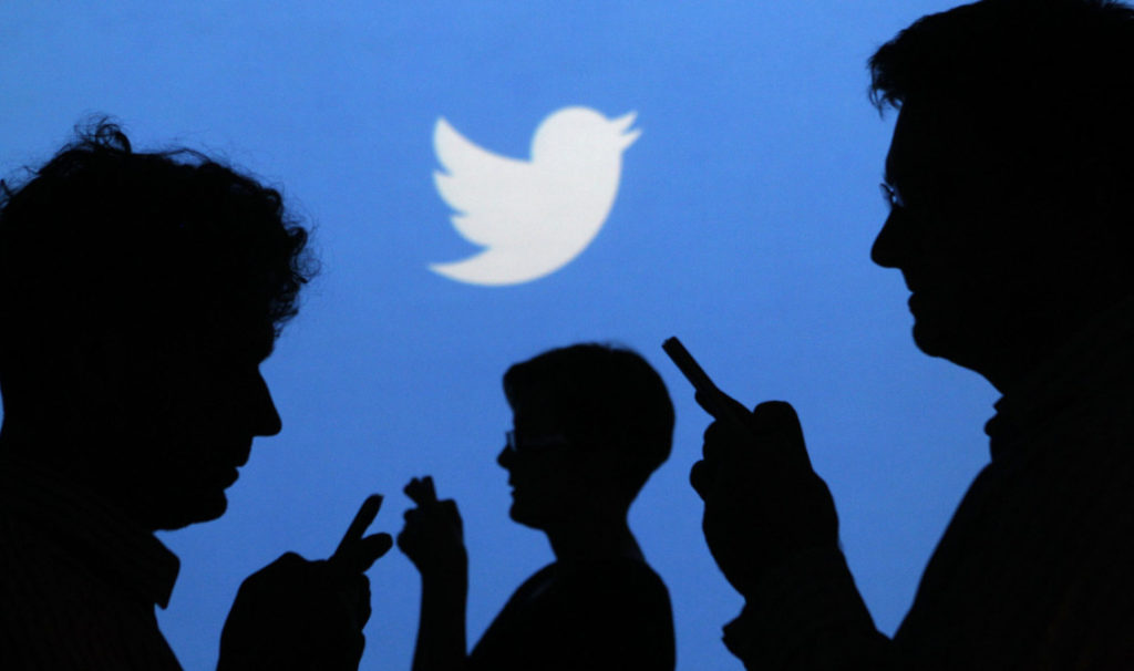 People holding mobile phones are silhouetted against a backdrop projected with the Twitter logo in Warsaw